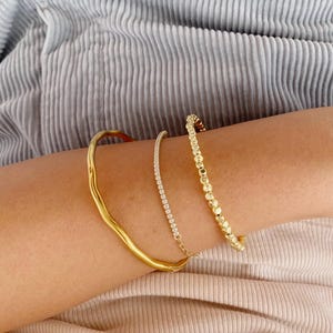 Può includere: Sono presentati tre braccialetti d'oro. Uno è una fascia strutturata e ondulata. Un altro è una delicata catena con piccole pietre trasparenti. Il terzo è un braccialetto di perline. I braccialetti sono impilati.