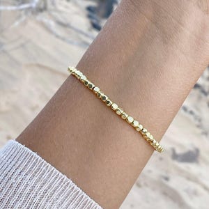Può includere: Un braccialetto dorato con perline dal design delicato. Il braccialetto è composto da piccole perline dorate, distanziate in modo uniforme. Il braccialetto è indossato su un polso, con una camicia bianca a maniche lunghe visibile.