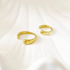 Könnte beinhalten: Zwei goldfarbene Ohrklemmen mit glatter, organischer Form. Das offene Design umschließt das Ohr und erzeugt einen modernen und minimalistischen Look. Der Schmuck wird auf einer weißen, strukturierten Oberfläche präsentiert.
