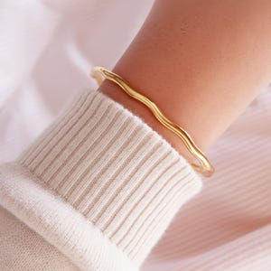 Può includere: Un bracciale rigido in metallo color oro caratterizzato da un design continuo, liscio e ondulato. Il bracciale presenta una superficie lucida e riflettente e una struttura semplice e aperta che avvolge il polso, con una finitura metallica.