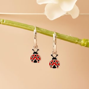 Pode incluir: Um par de brincos de argola em prata com pingentes em forma de joaninha. Cada pingente é esmaltado em vermelho brilhante com pontos pretos e uma cabeça preta. Os brincos estão suspensos num caule verde fino, destacando o acabamento em metal polido e o contraste vibrante do design de inseto vermelho e preto nas argolas de prata.