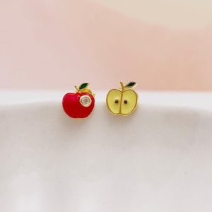 Può includere: Un paio di orecchini a forma di mela. Un orecchino è una mela rossa piena con una piccola gemma trasparente, l'altro è una fetta di mela gialla. Entrambi gli orecchini hanno una piccola foglia verde e perni color oro.
