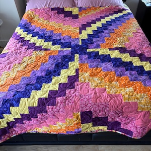 May include: A colorful quilt with a geometric pattern, featuring shades of pink, purple, yellow, and orange. The quilt is placed on a bed with pink pillows, showcasing a vibrant and handcrafted design. The quilt's dimensions are not visible.