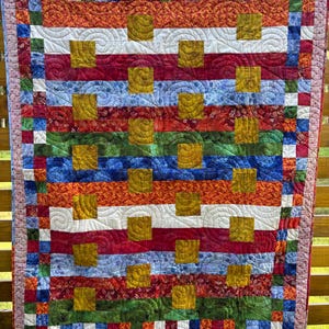 Peut inclure: Une courtepointe faite à la main aux couleurs vives, avec un motif patchwork. La courtepointe présente des rayures horizontales de différentes couleurs, dont le rouge, l'orange, le bleu, le vert et le blanc. Des carrés dorés sont espacés uniformément. La courtepointe est suspendue à une clôture en bois.
