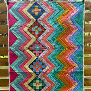 May include: A colorful handmade quilt with a zig-zag pattern in shades of pink, teal, orange, and yellow. The quilt features square patchwork designs in the center. The quilt is approximately 40 inches wide and 50 inches long.