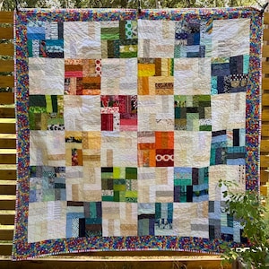 May include: A patchwork quilt with a geometric design. The quilt features squares of various fabrics in beige, green, orange, blue, and red. The border is a vibrant mix of colours. The quilt is hanging on a wooden fence. The quilt is approximately 150cm x 150cm.