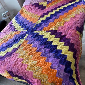 May include: A colorful quilt with a zig-zag pattern in shades of pink, purple, orange, and yellow. The quilt is on a bed with pink pillows. The quilt is handmade and has a quilted texture.