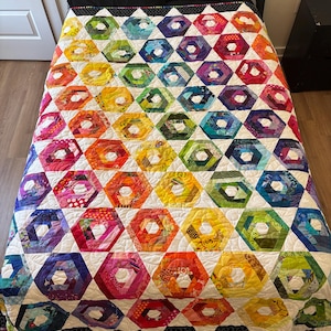 May include: A quilt featuring a repeating pattern of multicoloured hexagonal blocks arranged in a rainbow gradient. Each hexagon is constructed from various fabric scraps in shades of red, orange, yellow, green, blue, and purple, separated by white borders.