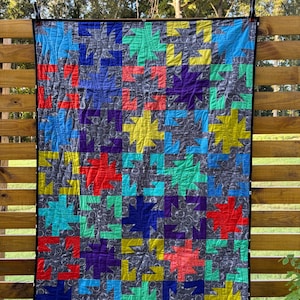 May include: A colorful quilt with a repeating starburst pattern in shades of blue, green, red, yellow, and purple. The quilt is displayed outdoors, hanging on a wooden fence. The background fabric is a dark gray with a subtle pattern.