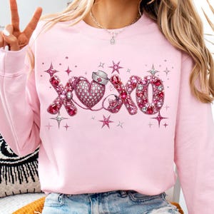 May include: A light pink sweatshirt featuring a sparkling "XOXO" design. The design includes a heart, a nurse's hat, and stars, all adorned with pink and clear rhinestones. The sweatshirt has long sleeves and a crew neck.