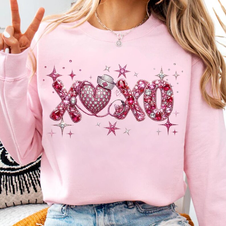 May include: A light pink sweatshirt featuring a sparkling "XOXO" design. The design includes a heart, a nurse's hat, and stars, all adorned with pink and clear rhinestones. The sweatshirt has long sleeves and a crew neck.
