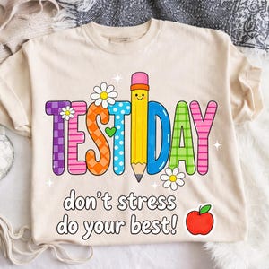 May include: Cream-colored t-shirt with a colorful "TEST DAY" graphic featuring a pencil, flowers, and the text "don't stress do your best!" A red apple is also included in the design. The shirt is made of a soft material.