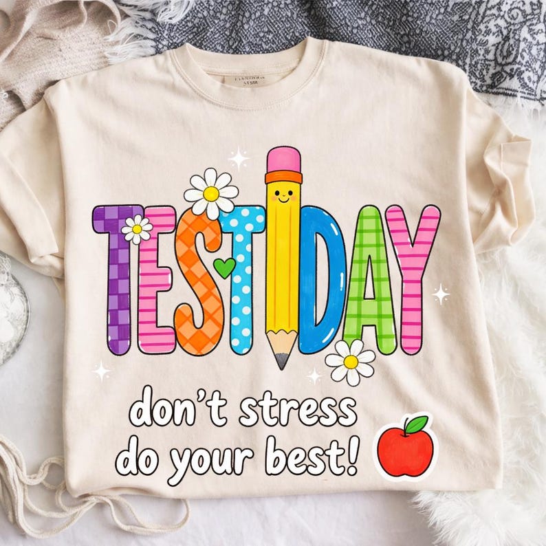 May include: Cream-colored t-shirt with a colorful "TEST DAY" graphic featuring a pencil, flowers, and the text "don't stress do your best!" A red apple is also included in the design. The shirt is made of a soft material.
