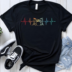 May include: Black t-shirt featuring a colorful heartbeat design with a drum set silhouette. The design transitions from red to yellow to blue. The shirt is paired with black and white sneakers and light blue denim shorts.