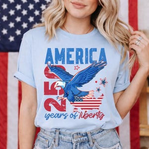 Könnte beinhalten: Hellblaues T-Shirt mit patriotischem Design. Das Shirt zeigt das Wort "AMERICA" in Blau, die Zahl "250" in Rot und eine Illustration eines Weißkopfseeadlers mit dem Text "years of liberty".