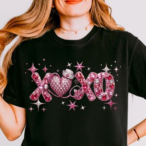 May include: Black t-shirt with a pink rhinestone design featuring the letters "XOXO", a heart, and a nurse's hat. The design is embellished with stars and sparkles. The shirt is a crew neck.