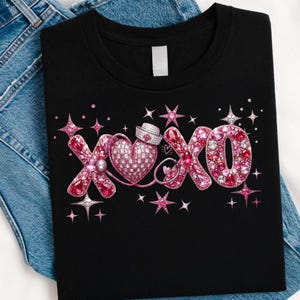 May include: Black t-shirt with a pink and white rhinestone design. The design features the letters "XO" with a heart, a nurse's hat, and stars. The shirt is folded on top of a pair of blue jeans.
