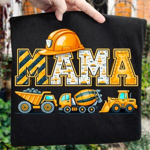 Puede incluir: Camiseta negra con la palabra "MAMA" en letras grandes de bloque amarillo y blanco con un tema de construcción. La letra "M" tiene rayas amarillas y negras. Debajo de la palabra hay ilustraciones de vehículos de construcción.