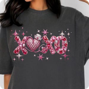 May include: Dark gray t-shirt with a pink and rhinestone design. The design features the letters "XOXO" with a heart, nurse's cap, and stars. The letters and heart are filled with pink rhinestones.