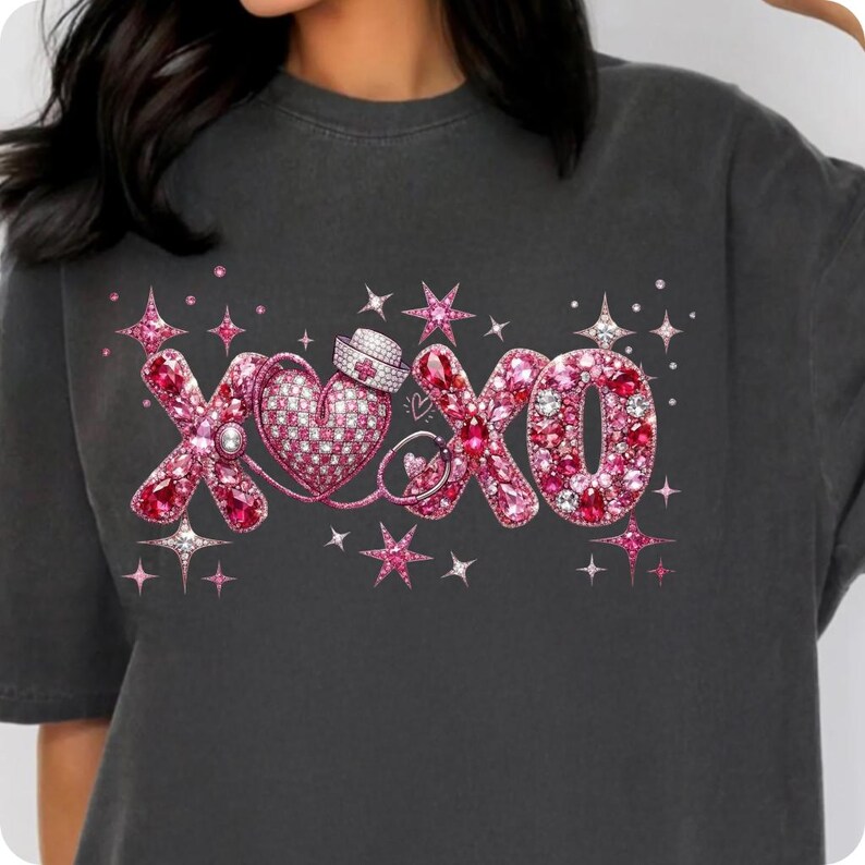 May include: Dark gray t-shirt with a pink and rhinestone design. The design features the letters "XOXO" with a heart, nurse's cap, and stars. The letters and heart are filled with pink rhinestones.