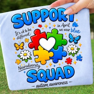 Op de afbeelding: Lichtblauw T-shirt met een kleurrijk grafisch ontwerp. Het ontwerp bevat puzzelstukjes in rood, groen, geel en blauw, met een wit hart in het midden. Tekst bevat "SUPPORT", "Neurodiversity SQUAD" en "autism awareness".