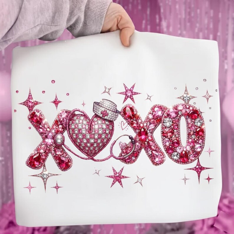 May include: White tote bag featuring the letters "XOXO" in pink gemstones and crystals, with a heart and nurse's cap design. Pink star accents surround the text. The bag is held by a person.