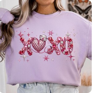 May include: A light purple sweatshirt with a rhinestone design. The design includes the letters "XOXO", a heart, a stethoscope, and stars. The letters and heart are embellished with pink and red rhinestones. The sweatshirt is a casual style.