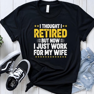 Op de afbeelding: Zwart t-shirt met de tekst "I THOUGHT I RETIRED BUT NOW I JUST WORK FOR MY WIFE" in geel en wit. Het shirt wordt gecombineerd met zwart-witte sneakers en een blauwe jeans.