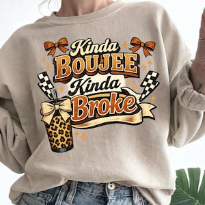 May include: Beige sweatshirt with the text "Kinda Boujee Kinda Broke" in brown and orange lettering. The design includes a leopard print cup with a bow, orange bows, and checkered flags.