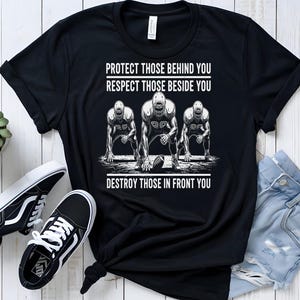 May include: Black t-shirt with the text "PROTECT THOSE BEHIND YOU RESPECT THOSE BESIDE YOU DESTROY THOSE IN FRONT YOU" and a graphic of three football players. Black and white trainers and a small potted plant are also in the image.