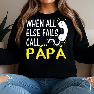May include: Black sweatshirt with the text "WHEN ALL ELSE FAILS CALL... PAPA" in white and yellow. The design includes a white telephone graphic. The sweatshirt is worn by a person sitting on a couch.