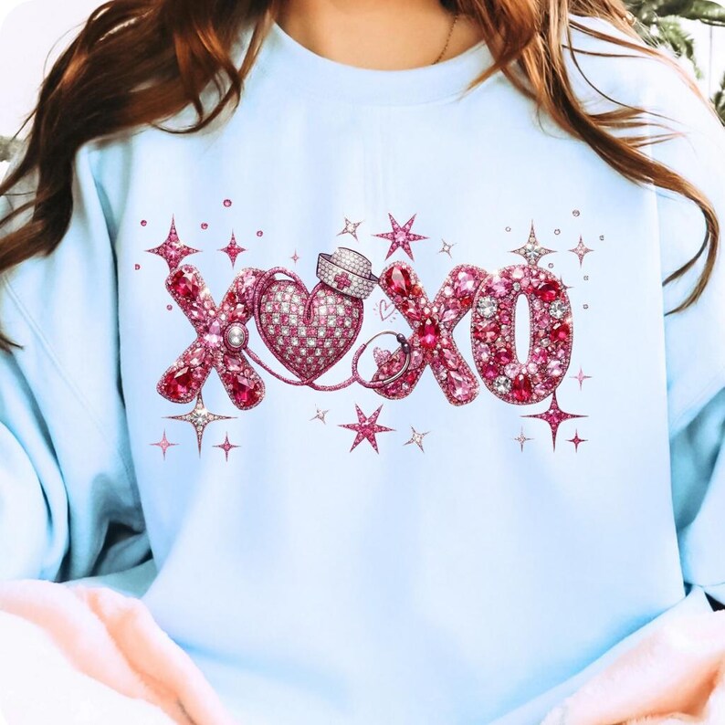 May include: Light blue sweatshirt featuring a pink and red rhinestone design of the letters "XOXO" with a heart and stethoscope. The design includes stars and sparkles, creating a sparkling effect.