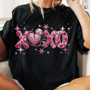 May include: Black t-shirt featuring a pink XOXO design with a heart and nurse's hat. The letters and heart are embellished with pink rhinestones and surrounded by star accents. A cute and romantic design.