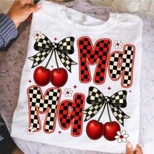 May include: White t-shirt with the word "Mad" in a checkered pattern with red accents. The design includes cherries, bows, and small flowers. The shirt is held up by a person.
