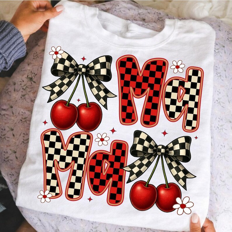 May include: White t-shirt with the word "Mad" in a checkered pattern with red accents. The design includes cherries, bows, and small flowers. The shirt is held up by a person.