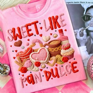 May include: Pink t-shirt with the words "SWEET LIKE PAN DULCE" in raised, textured lettering. The design includes embroidered hearts, pastries, a coffee cup, and a cupcake. The overall theme is a celebration of sweets and baked goods.