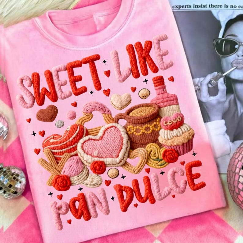 May include: Pink t-shirt with the words "SWEET LIKE PAN DULCE" in raised, textured lettering. The design includes embroidered hearts, pastries, a coffee cup, and a cupcake. The overall theme is a celebration of sweets and baked goods.