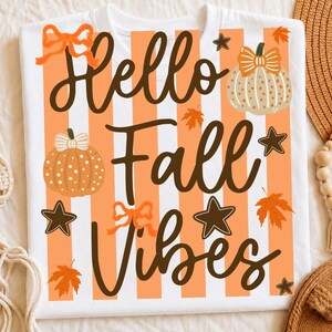 Op de afbeelding: Een wit t-shirt met een afbeelding van oranje verticale strepen en de donkerbruine tekst Hello Fall Vibes. Het ontwerp bevat illustraties van twee pompoenen, oranje esdoornbladeren, donkerbruine sterren en twee oranje strikken.
