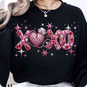 May include: Black sweatshirt with a pink and red rhinestone design. The design features the letters "XOXO" with a heart, stars, and a nurse's hat. The letters and heart are embellished with rhinestones, creating a sparkling effect.