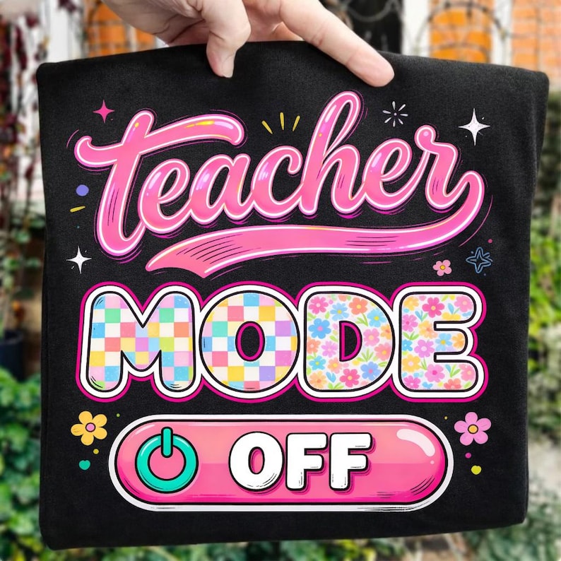 May include: Black t-shirt with the words "Teacher MODE OFF" in a colorful, playful design. The word "Teacher" is in pink script, "MODE" has a checkered pattern, and "OFF" is in a pink button shape.