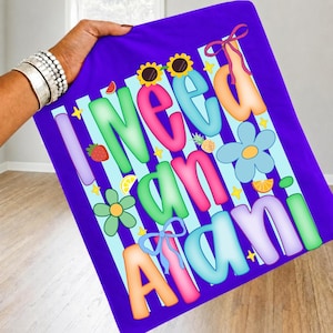 May include: A purple t-shirt with the phrase "I Need An Alani" in colorful bubble letters. The design includes sunflowers, flowers, fruit, and bows. The shirt is held up by a person.