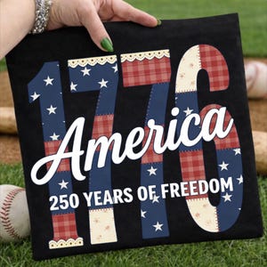 Op de afbeelding: Zwart T-shirt met het woord "America" in witte letters. De cijfers "1776" zijn in een patchwork-ontwerp van rood geruit, blauw met witte sterren en crèmekleurige bloempatronen. De tekst "250 YEARS OF FREEDOM" staat eronder.
