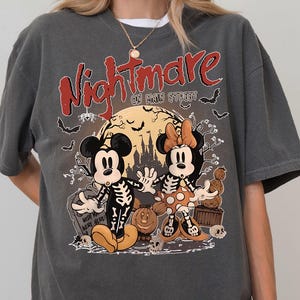Könnte beinhalten: Ein anthrazitfarbenes Kurzarm-T-Shirt mit einem Grafikdruck und dem Text Nightmare on Main Street. Das Design zeigt zwei Zeichentrickmäuse in Skelettkostümen, einen großen gelben Mond, eine Schloss-Silhouette, Fledermäuse und Kürbisse.
