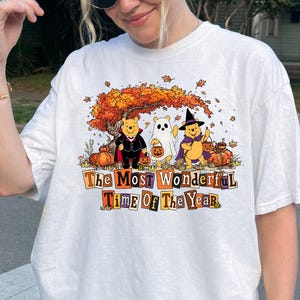 Könnte beinhalten: Ein weißes Kurzarm-T-Shirt mit einer Grafik von Winnie Puuh-Figuren in Halloween-Kostümen unter einem Herbstbaum. Der Text lautet The Most Wonderful Time Of The Year, umgeben von Kürbissen und herbstlichen Blättern auf dem Shirt.