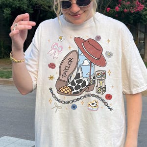 Pu&ograve; includere: Una t-shirt a maniche corte color bianco sporco con una stampa grafica raffigurante uno stivale da cowboy maculato con il nome TAYLOR, un cappello da cowboy marrone, blocchi con la scritta BTS, una catenella di perline e fiori e stelle.