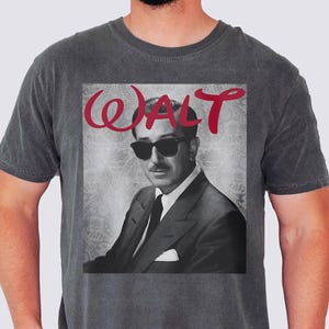 Könnte beinhalten: Dunkelgraues T-Shirt mit einem Schwarz-Weiß-Bild eines Mannes mit Sonnenbrille und Anzug. Das Wort "WALT" ist in roter Schreibschrift über dem Bild geschrieben. Das T-Shirt ist einfarbig und hat kurze Ärmel.