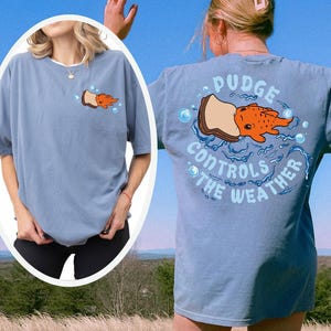 K&ouml;nnte beinhalten: Hellblaues T-Shirt mit einer Grafik eines Cartoon-Fisches auf einem Sandwich, mit dem Text "Pudge Controls the Weather". Das Shirt hat eine lockere Passform und kurze &Auml;rmel. Das Design ist auf der Vorder- und R&uuml;ckseite aufgedruckt.