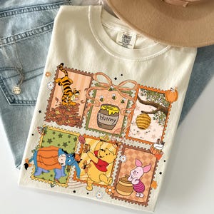 Könnte beinhalten: Ein cremefarbenes T-Shirt mit einem Grafikdruck aus sechs rechteckigen Rahmen im Briefmarkenstil. Die Rahmen enthalten Illustrationen von Tigger im Laub, einem Honigtopf mit der Aufschrift Hunny, einem Bienenstock, I-Aah, Winnie Puuh und Ferkel.