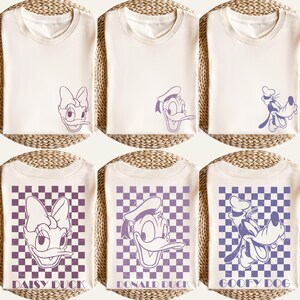 Puede incluir: Camisetas de color crema con dise&ntilde;os de personajes de dibujos animados. Los dise&ntilde;os incluyen a Daisy, Donald y Goofy. Algunas camisetas tienen un fondo de tablero de ajedrez p&uacute;rpura con los nombres de los personajes. Las camisetas est&aacute;n dobladas y expuestas sobre una superficie tejida.