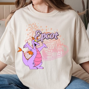 May include: Cream-colored t-shirt featuring a purple dragon character with orange wings and a pink belly. The word "Epcot" is written above the dragon in a stylized font, with a pink outline of a theme park in the background.